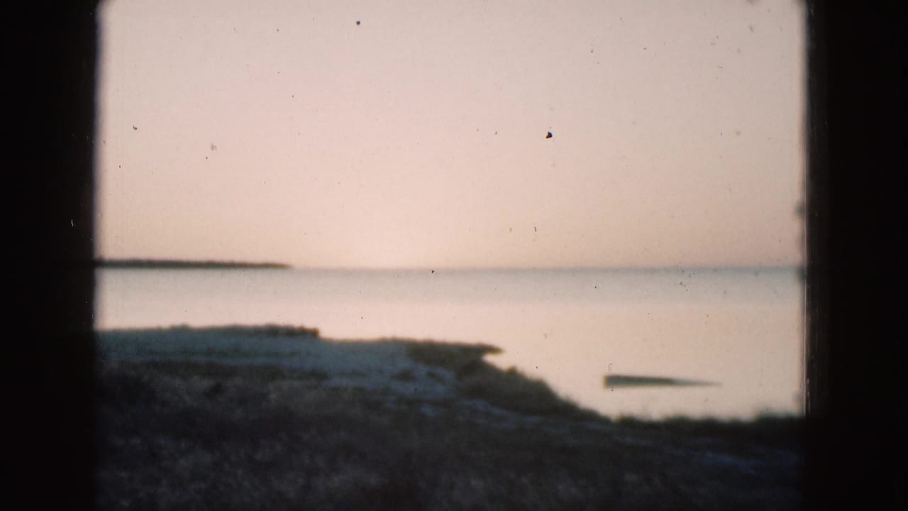 1975 Florida 8mm frame showing a quiet shoreline and calm water under a pale pink-blue sky, bordered by dark film edges.