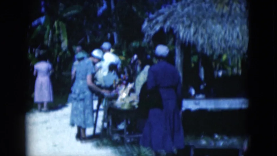 1975 Florida 8mm frame of adult visitors in dresses and hats gathered near a shaded zoo or wildlife viewing area.