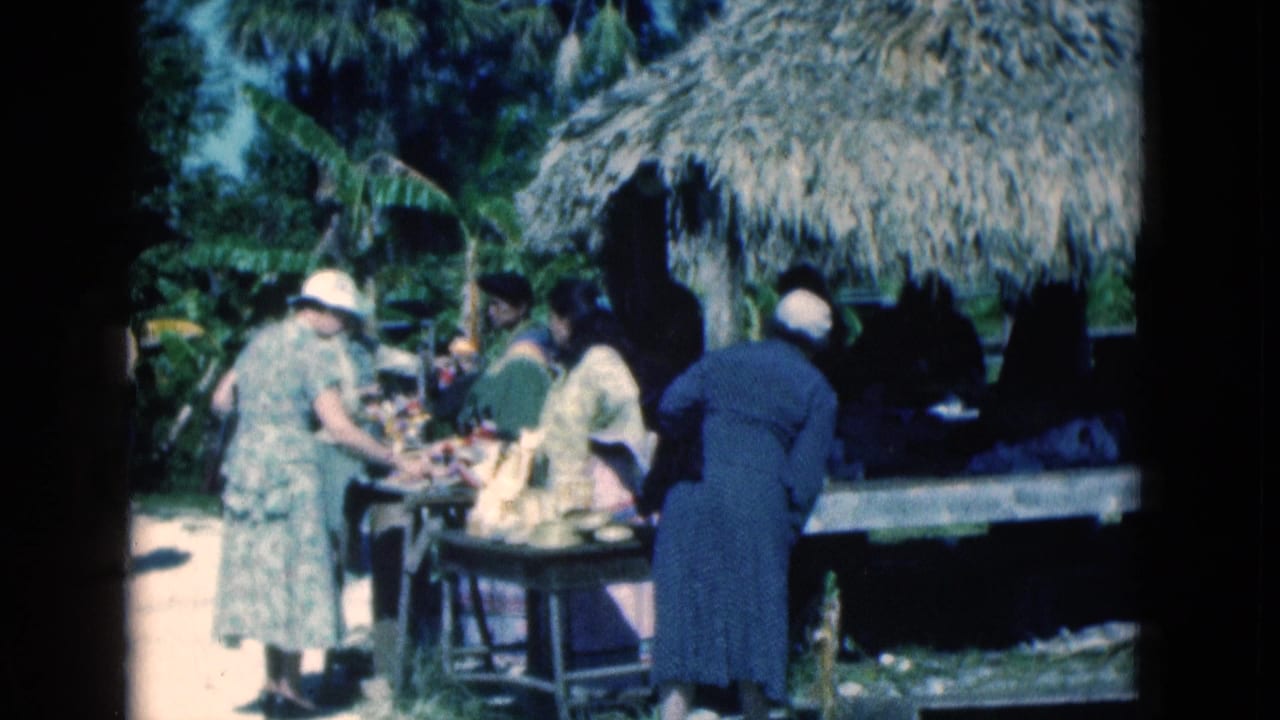 Adults gather around a table or rail under a thatched shelter in a tropical zoo setting.