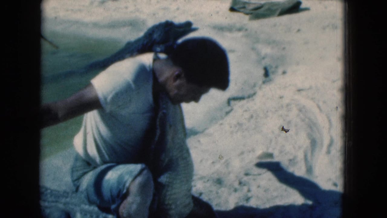 Man in a white shirt crouches over a large crocodile-like reptile on pale sand.