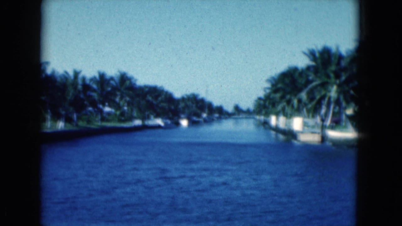1975 Florida 8mm frame showing a calm canal bordered by palms, seawalls, and distant waterfront residences.