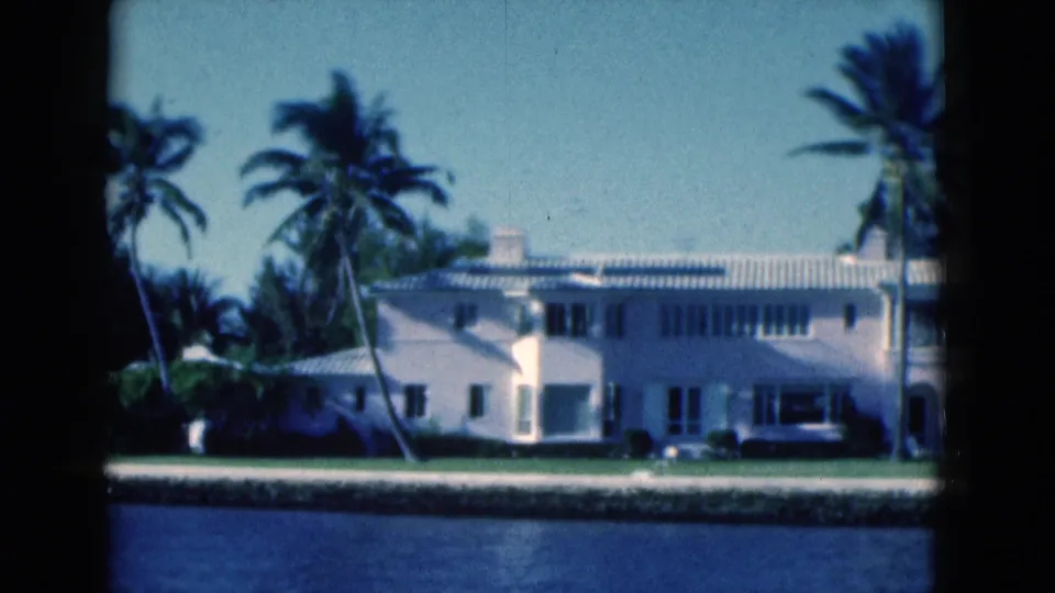 1975 Florida home-movie frame of a white canal-side house, palms, lawn, and seawall viewed from across the water.