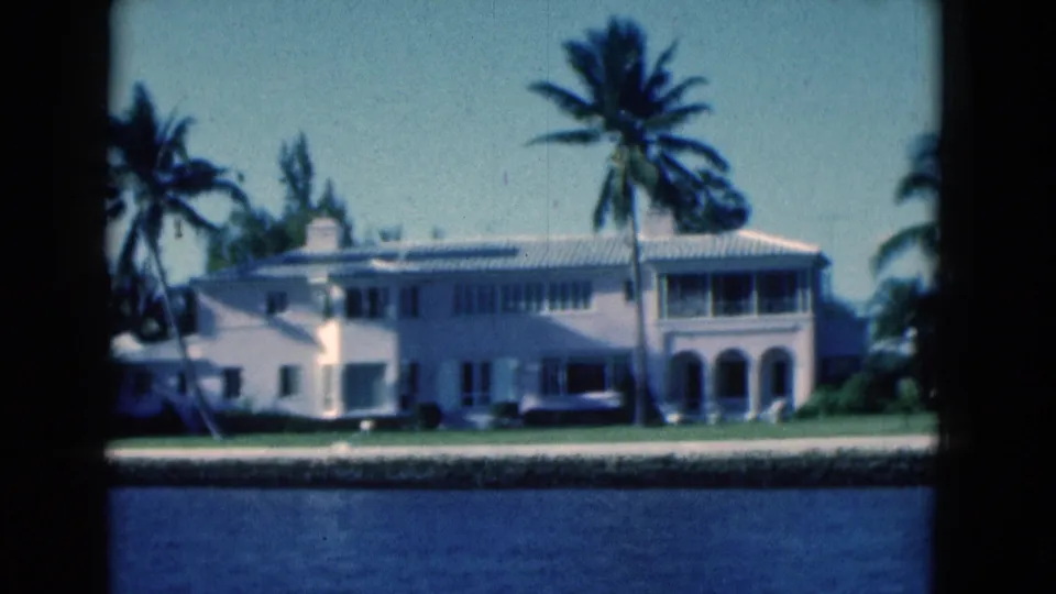 1975 Florida 8mm view of a canal-front house with arched porch details, palms, lawn, and water in the foreground.
