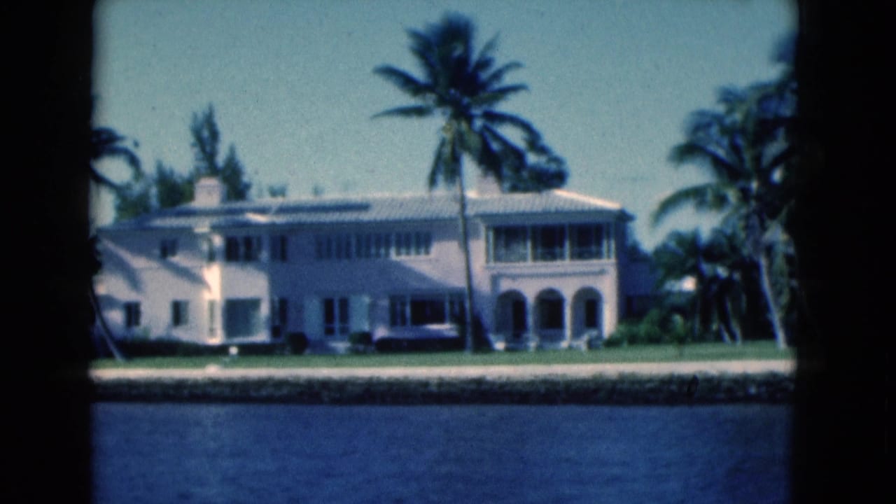 1975 Florida 8mm frame showing a white riverside villa, palm trees, seawall, and blue canal water.