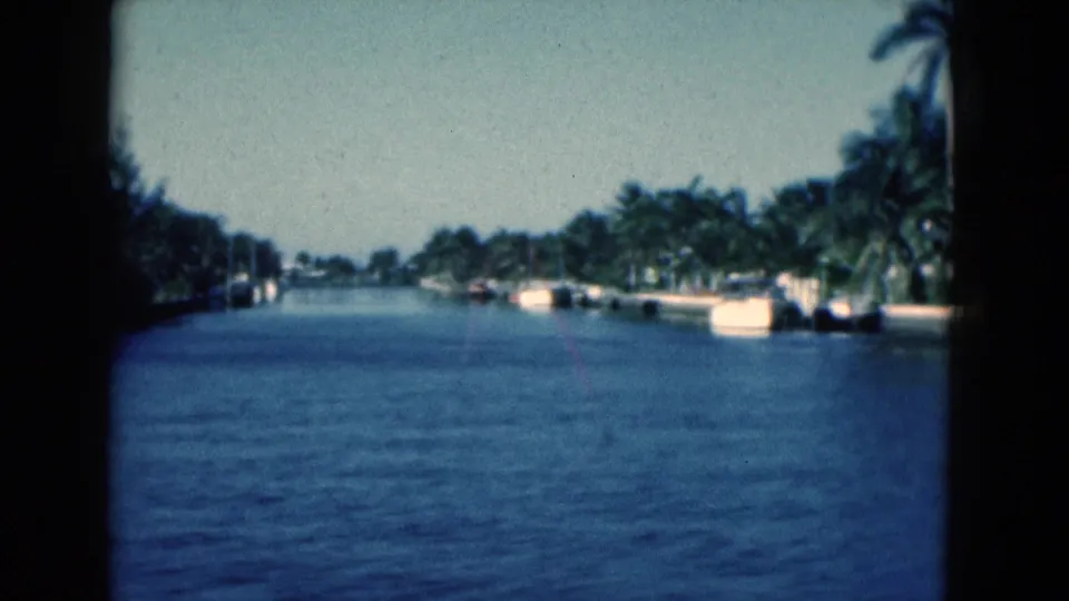 1975 Florida 8mm frame looking along a blue canal with palms, docks, small boats, and low waterfront structures.