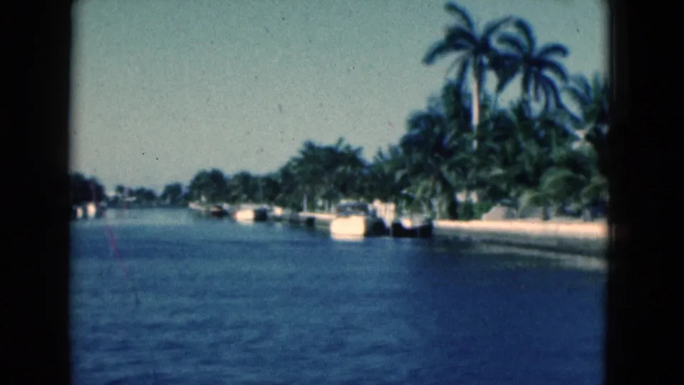 1975 Florida canal scene on 8mm film, showing palms, docked boats, seawalls, and sunlit waterfront greenery.