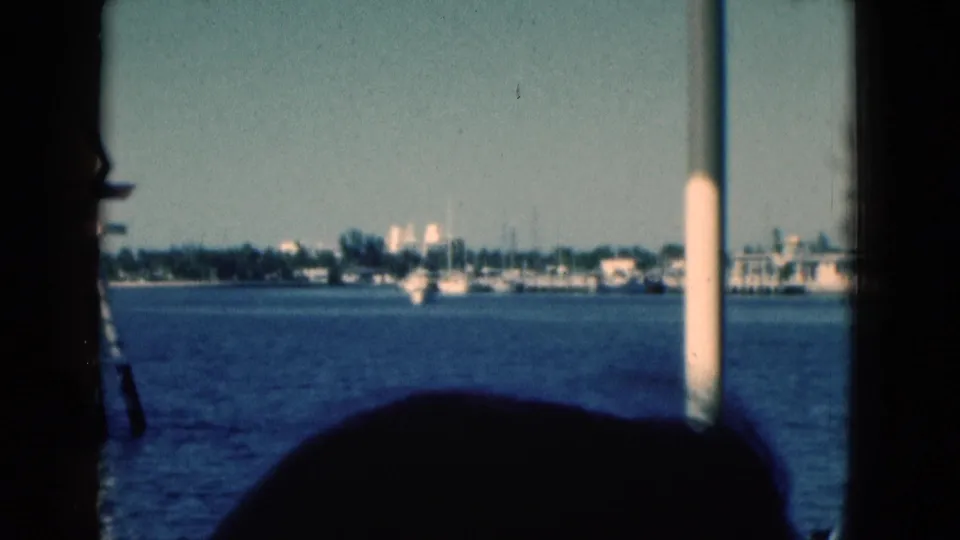 1975 Florida 8mm boat-view frame showing blue water, distant marina boats, shoreline buildings, and dark foreground silhouettes.