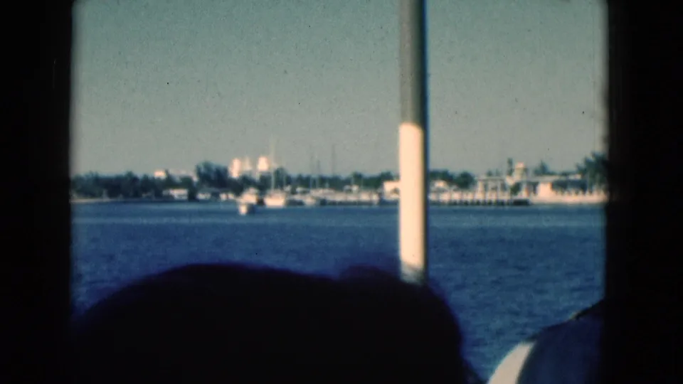 1975 Florida coastal waterway frame with marina boats, low buildings, a vertical boat post, and dark passenger or boat silhouettes.