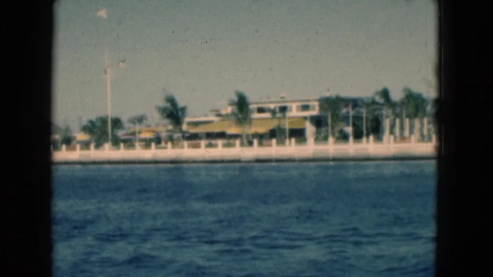 1975 Florida 8mm frame with a palm-lined waterfront, white seawall and light-colored building viewed from moving water.