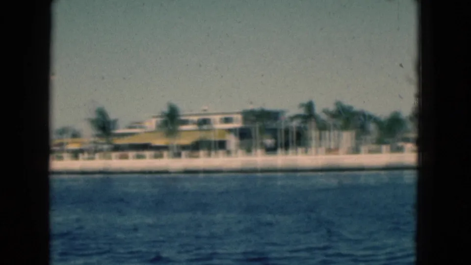 1975 Florida 8mm frame showing choppy blue water in front of a white seawall, palm trees and a low yellow-roofed building.