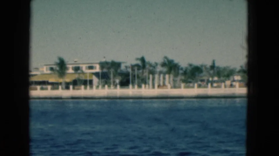 1975 Florida 8mm frame with dark gate borders around a waterfront view of blue water, palms, a seawall and low building.