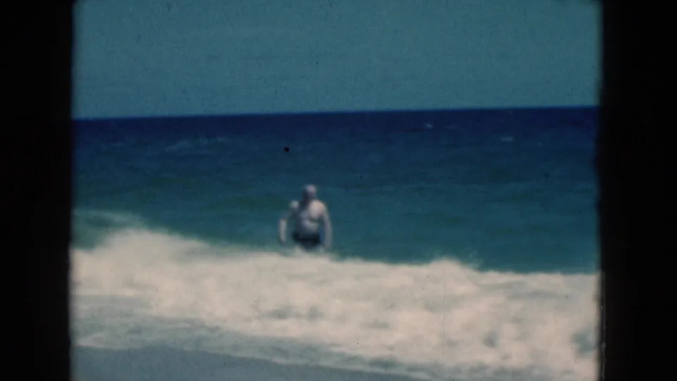 1975 Florida 8mm frame showing a man in a swim cap standing in breaking ocean waves under a blue sky.