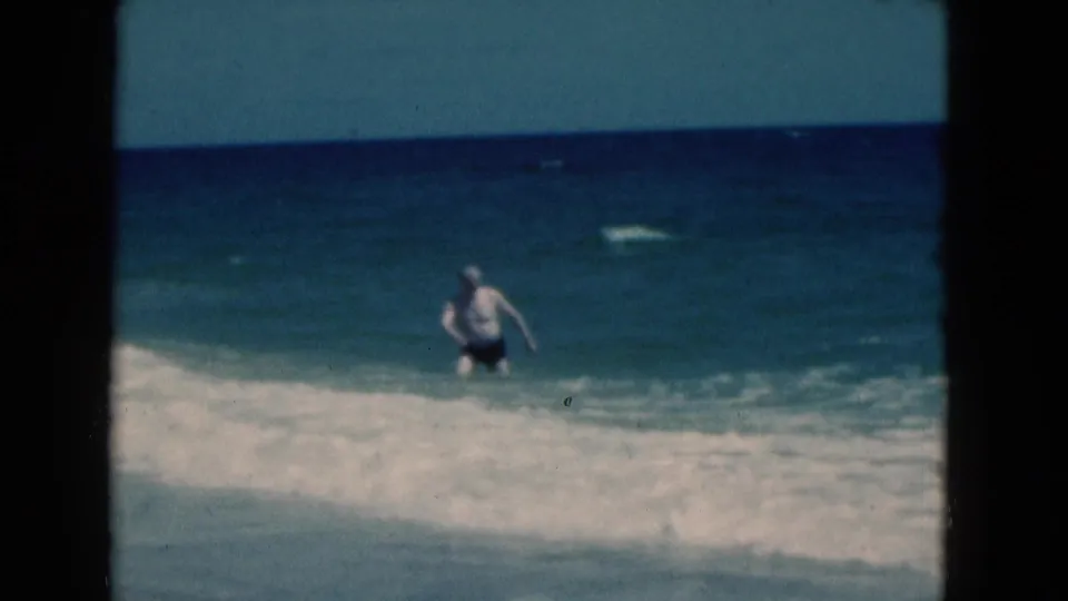 1975 Florida 8mm frame showing a man in dark swim trunks stepping through shallow whitewater near the beach.
