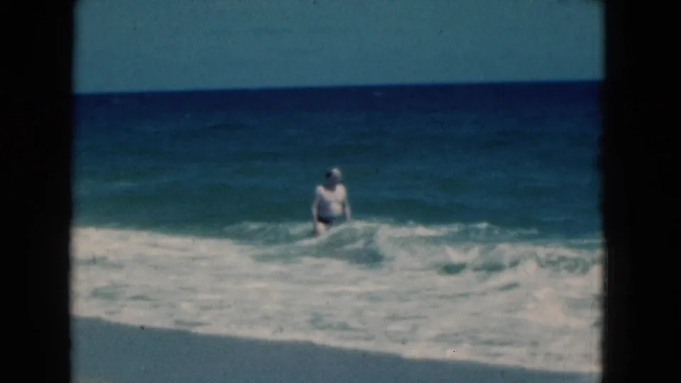 1975 Florida home-movie frame with a man standing in rolling surf as white foam moves toward shore.