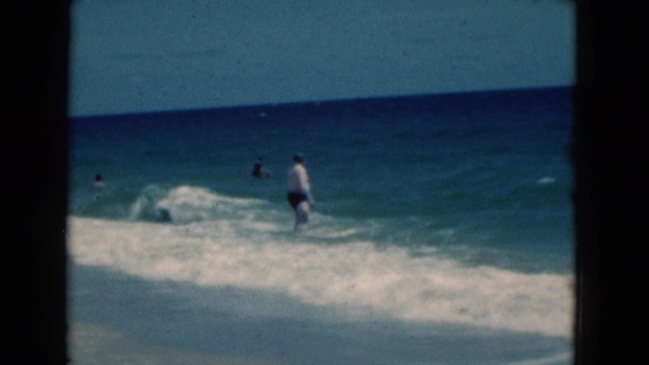 1975 Florida 8mm frame showing a man walking through shallow ocean waves with white surf and distant swimmers in the water.