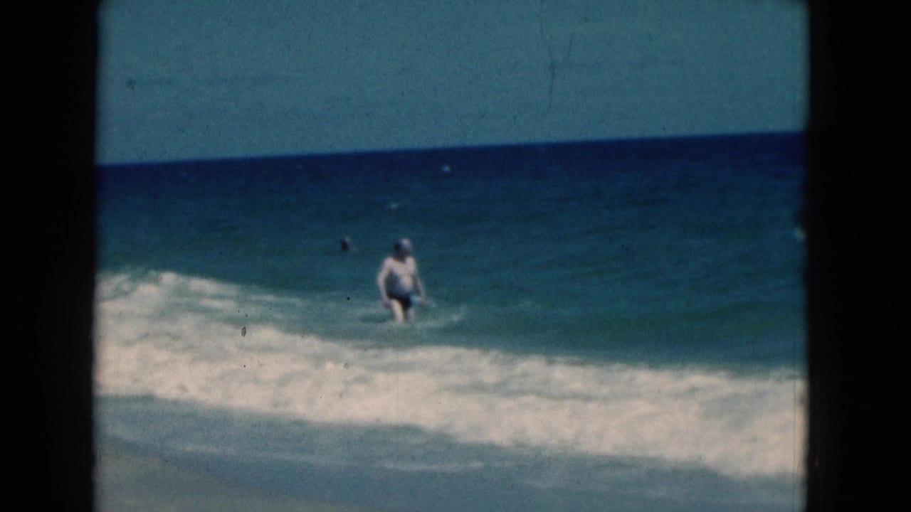 Man stands in the ocean as a low wave breaks in front of him.