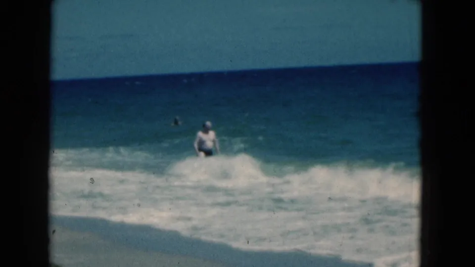 Soft 8mm frame from a 1975 Florida beach showing a man partly surrounded by foam as ocean waves break near shore.