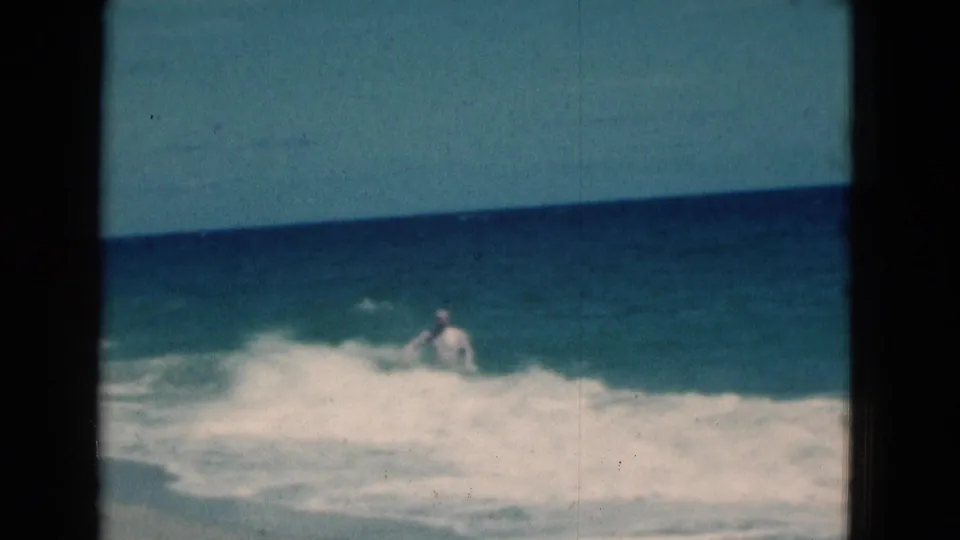 1975 Florida 8mm beach frame with the man partly hidden behind foamy surf under a blue ocean horizon.