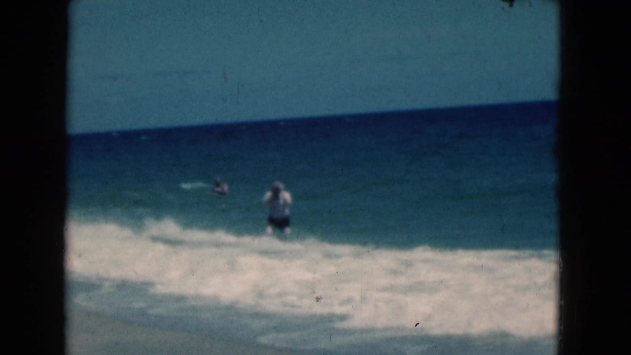 Man in a white shirt wading in ocean surf with a distant swimmer beyond him.