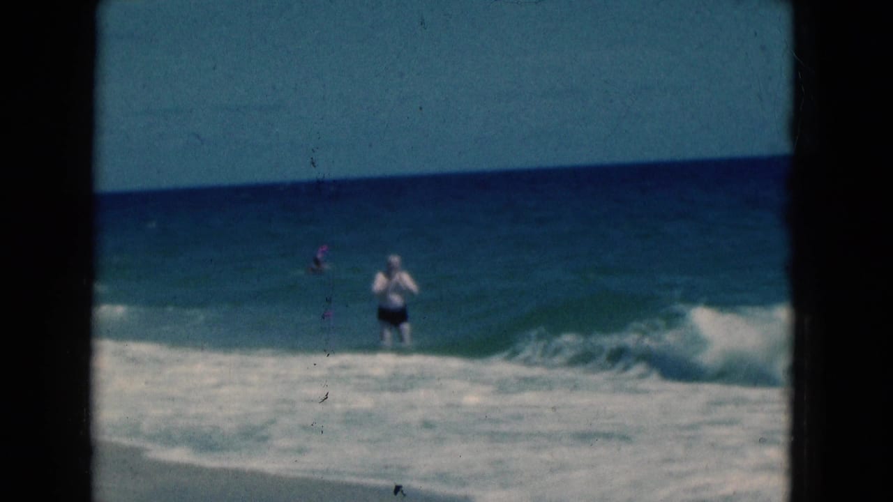1975 Florida beach frame showing a man in dark shorts and a white shirt amid rolling ocean waves under a blue horizon.