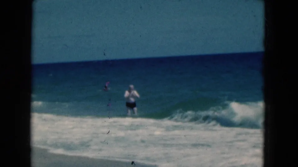 1975 Florida beach frame showing a man in dark shorts and a white shirt amid rolling ocean waves under a blue horizon.