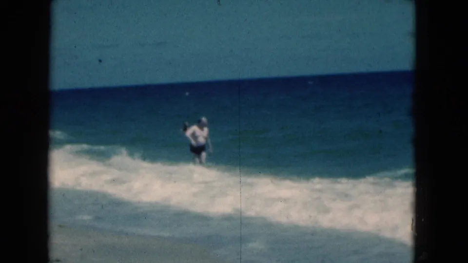 1975 Florida 8mm frame with a man moving through white surf close to the sand, filmed from a distant beach vantage.