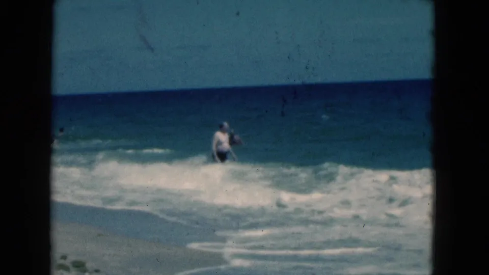 1975 Florida beach frame of a man in a white shirt standing amid foamy waves, with blue ocean and sky in the background.