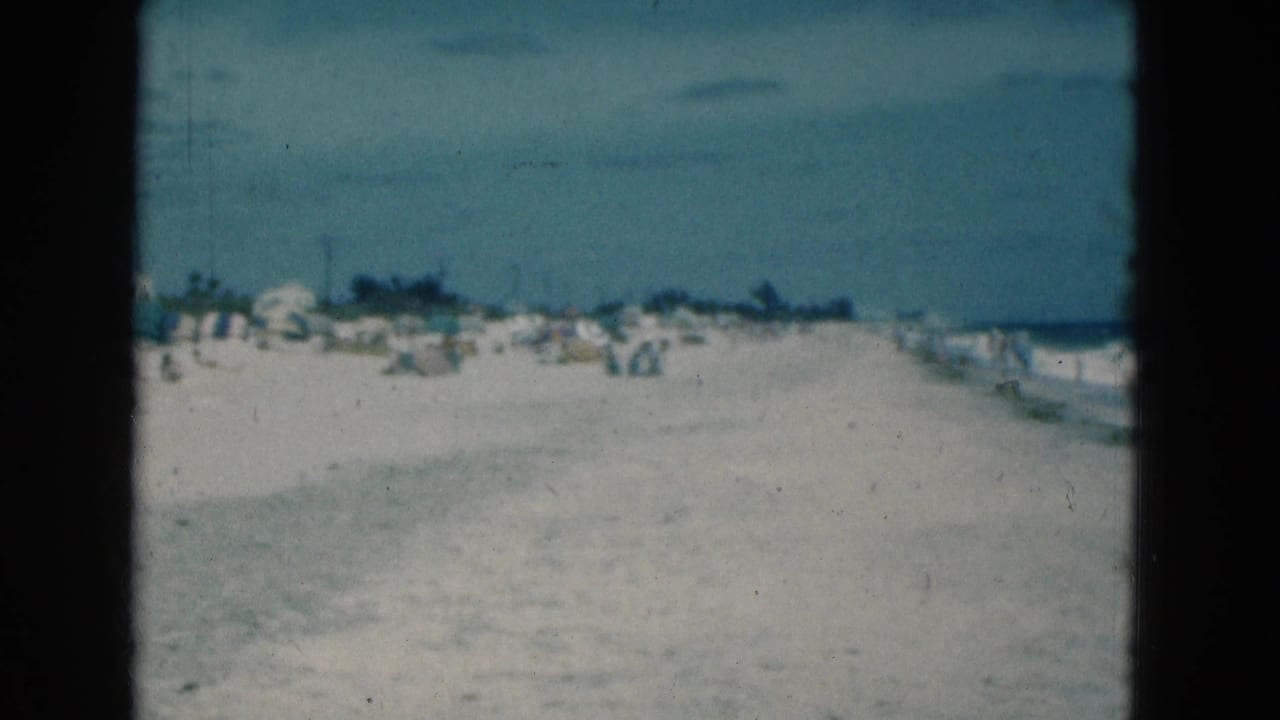 1975 Florida beach scene with scattered umbrellas, small shoreline figures, pale sand, and surf visible at frame right.