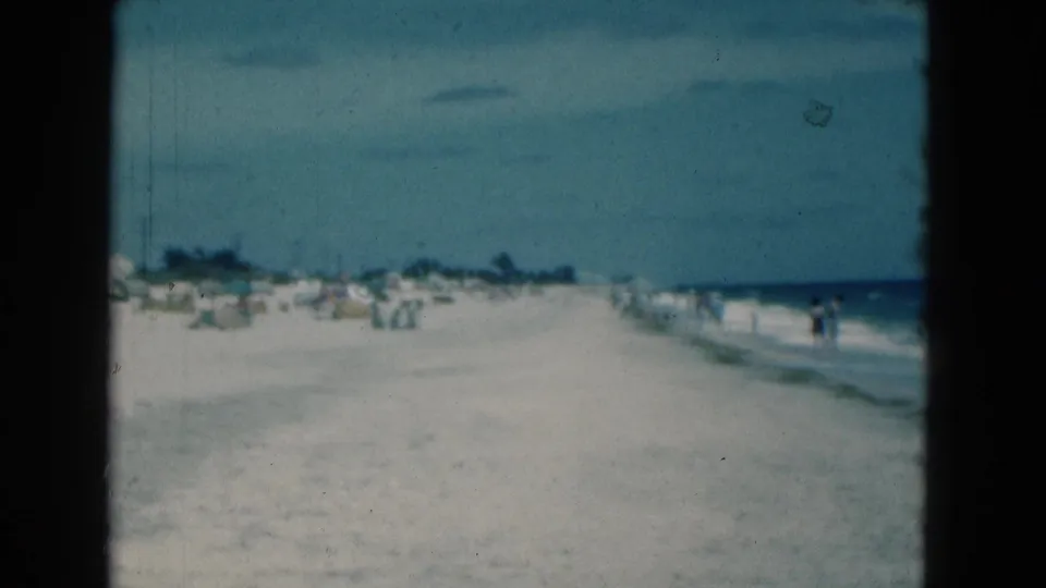 Soft 1975 Florida shoreline view with beachgoers in the distance, umbrellas on sand, and ocean water along the right side.