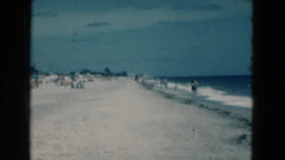 1975 Florida 8mm frame showing a broad beach, small figures by the water, umbrellas, surf, and faded blue sky.