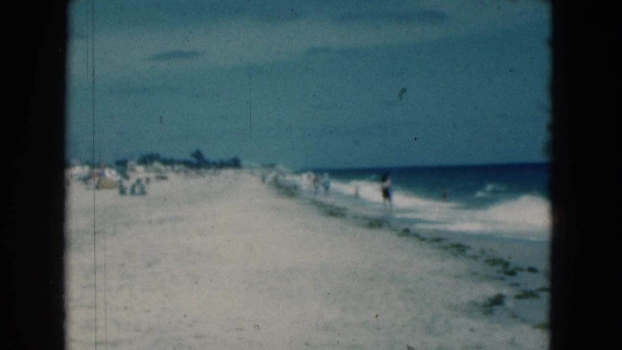 1975 Florida 8mm view of a white sand beach with distant beachgoers, blue ocean, and gentle surf under open sky.