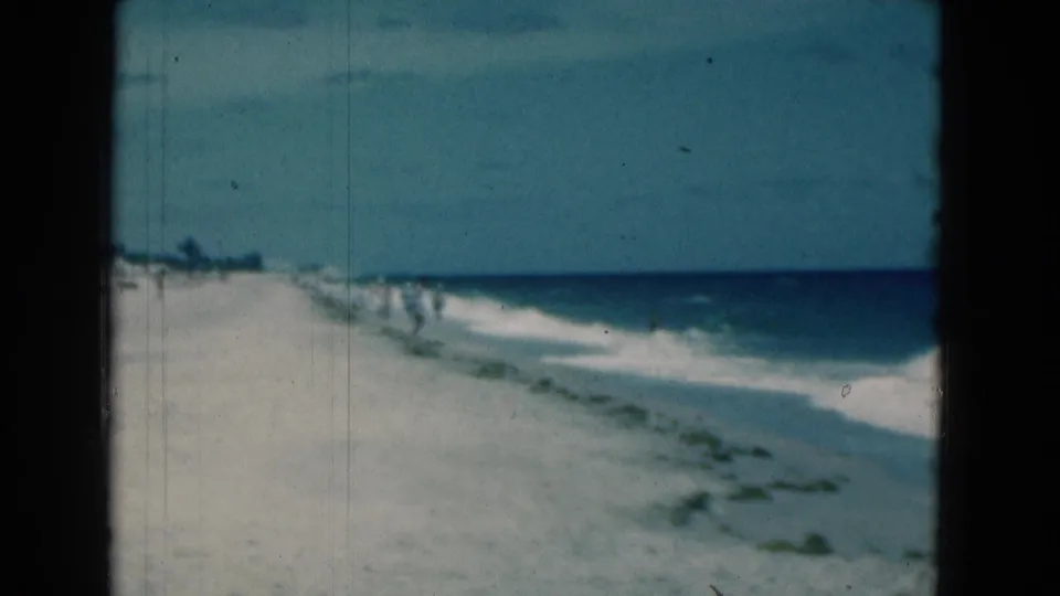 Soft 1975 Florida shoreline frame with white sand, blue surf, distant beachgoers, and visible film grain from 8mm home-movie footage.