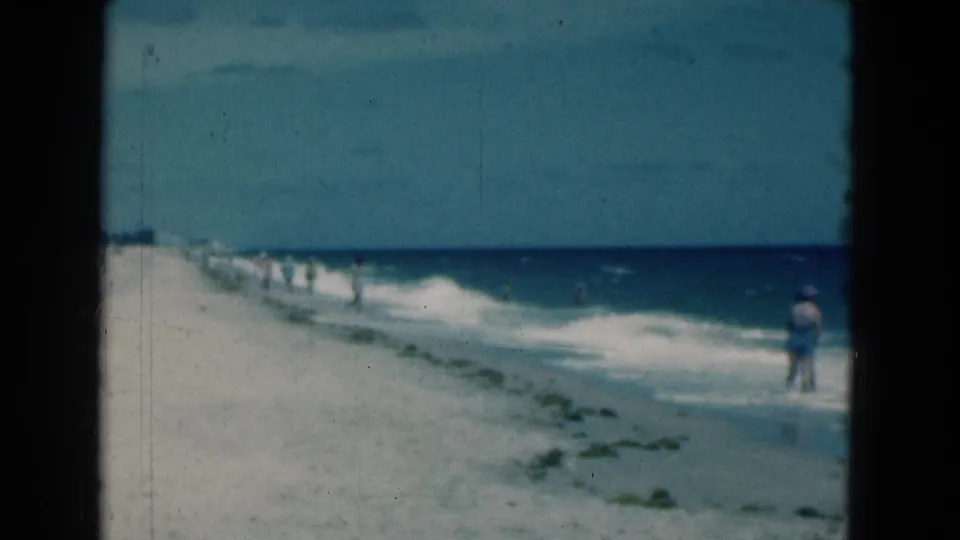 1975 Florida 8mm beach frame with one person standing near the waves and more beachgoers farther down the shoreline.