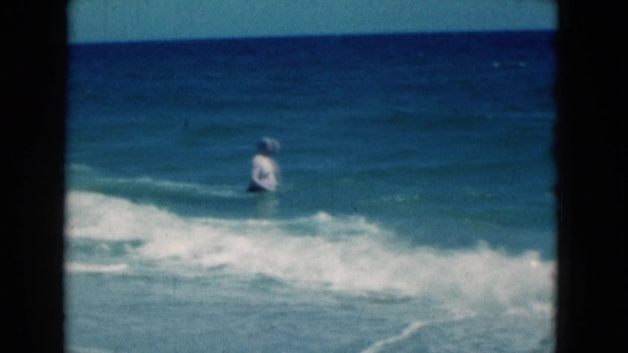 In 1975 Florida beach footage, an adult man in a white swimsuit stands in blue surf with a low wave rolling in.