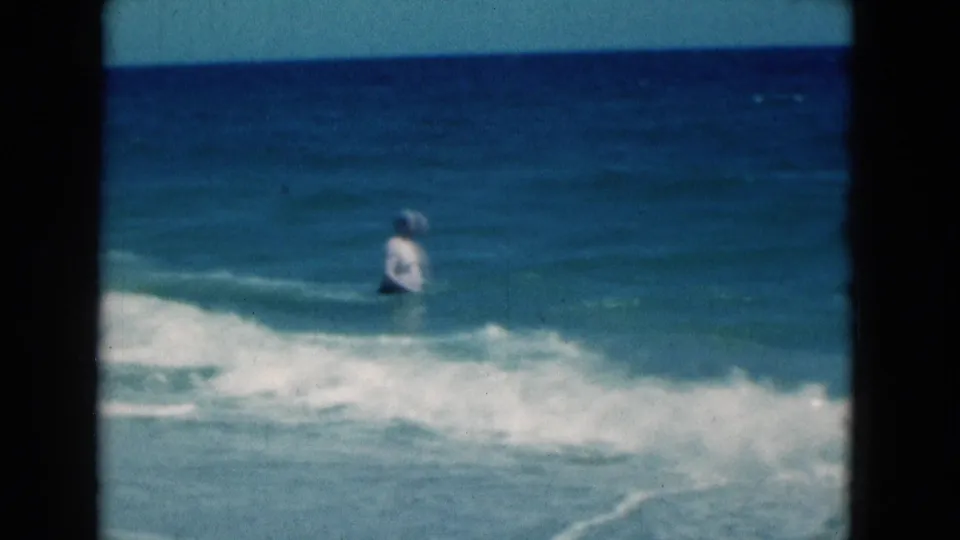 In 1975 Florida beach footage, an adult man in a white swimsuit stands in blue surf with a low wave rolling in.
