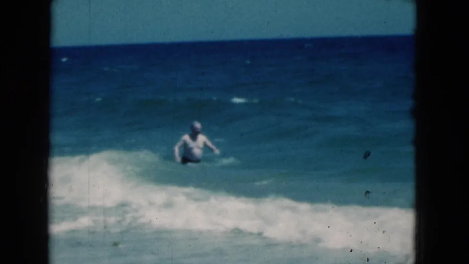 At the end of the 1975 Florida beach clip, the man faces the open water while a gentle wave lifts around his waist.