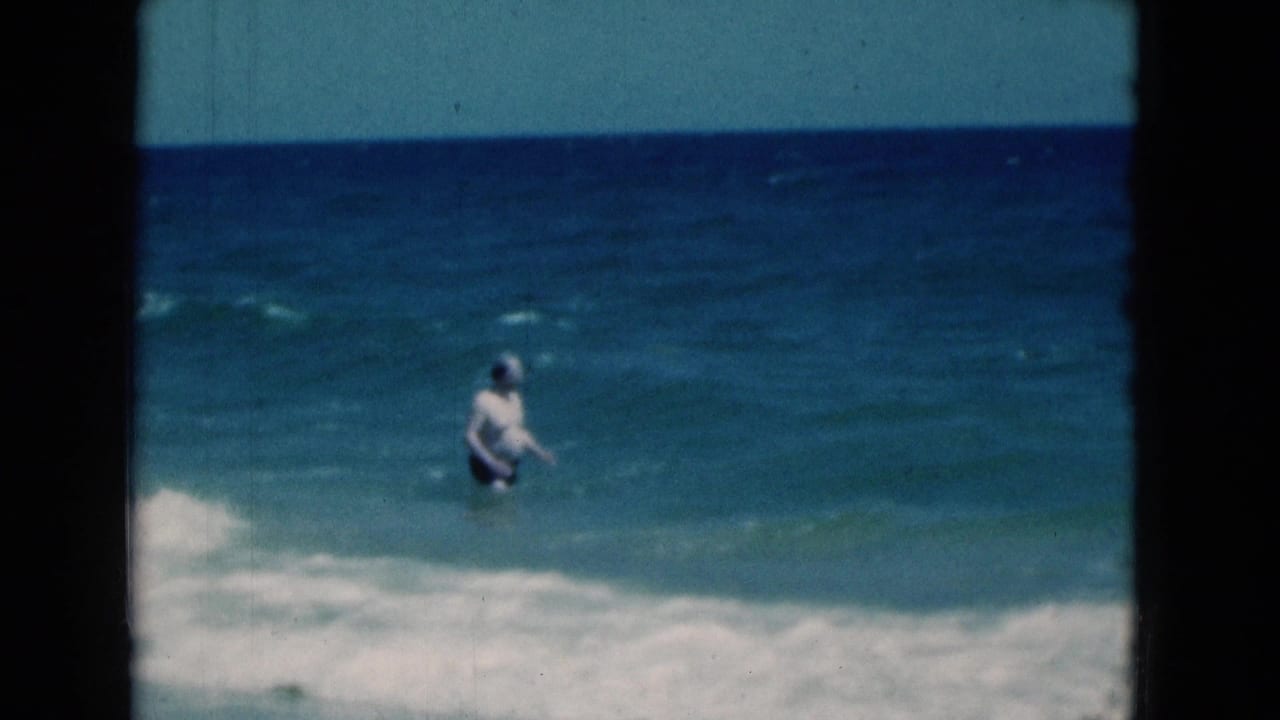 In 1975 Florida beach footage, a man in swim trunks wades in blue ocean water as small waves approach the shore.