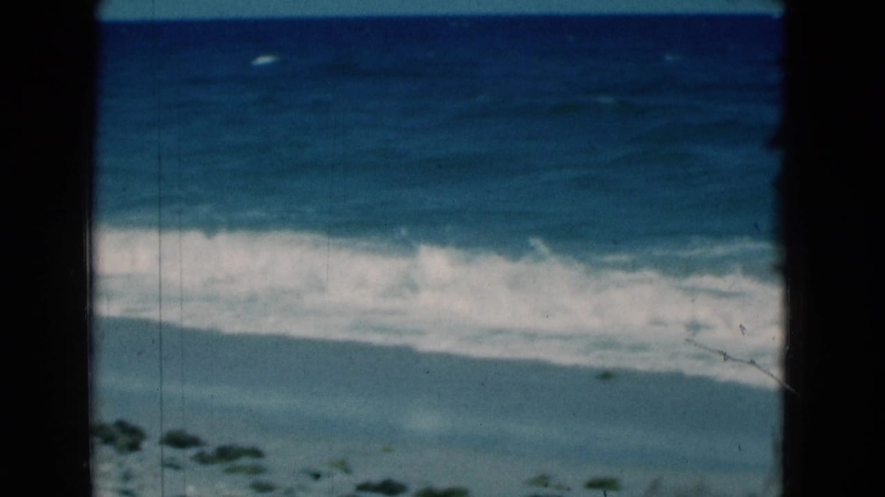 White surf line crosses blue water and sand within a dark film gate frame.