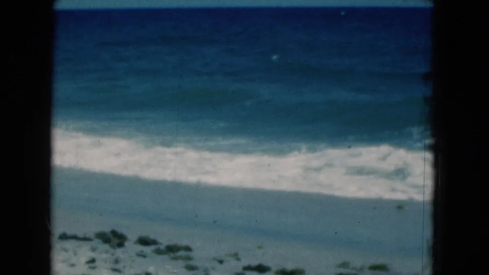 1975 Florida 8mm still showing foamy ocean water reaching a quiet beach with low vegetation visible near the bottom.