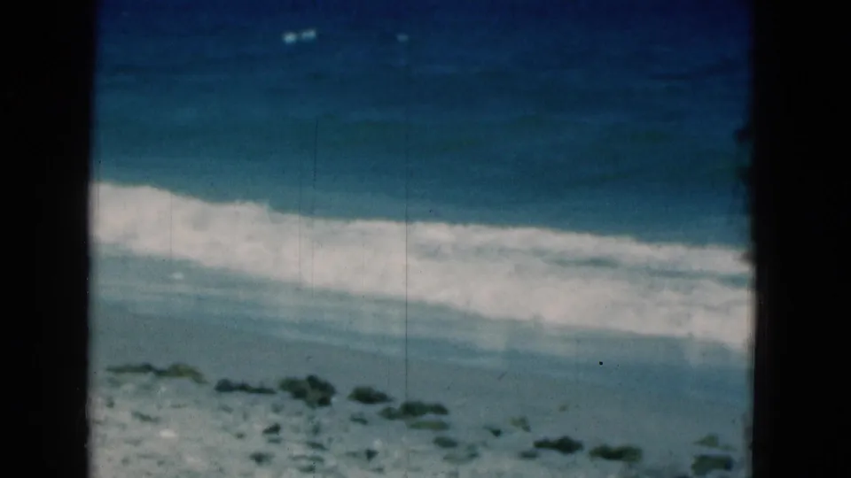 1975 Florida coastal 8mm frame with a diagonal surf line, blue water, pale sand, and no visible people or structures.