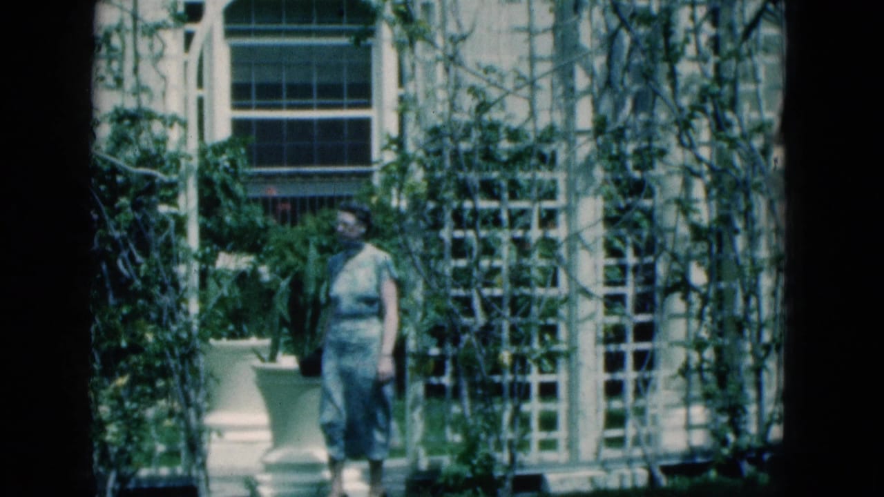 In 1975 Florida home-movie footage, a woman stands near vine-covered white trellises and a tall window in bright sun.