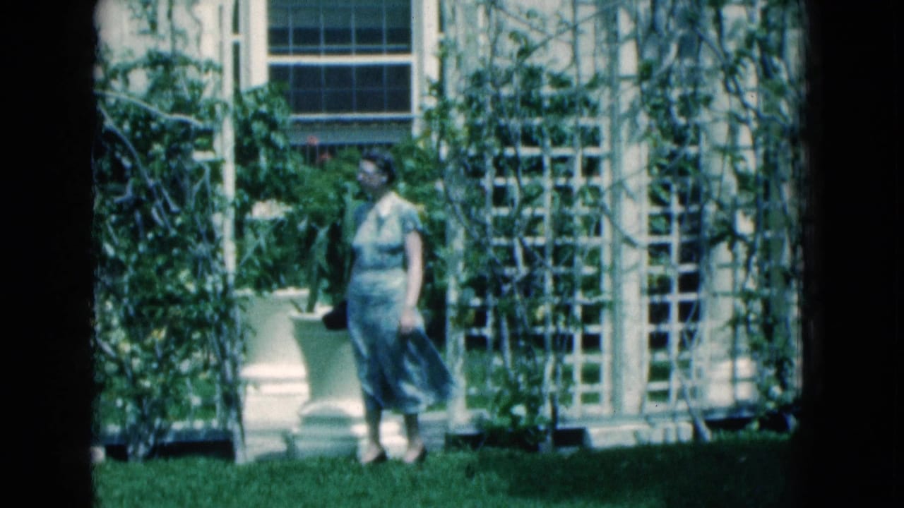 A woman in a light dress stands on a green lawn beside planters, vines, and white trelliswork in Florida, 1975.