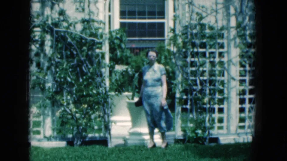 A woman faces the camera in a sunlit 1975 Florida garden, with white trellises, climbing vines, and a window behind her.