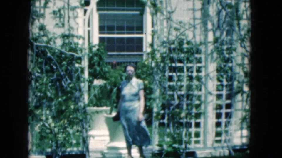 A woman stands still near a large planter and vine-covered white trellis in 1975 Florida home-movie footage.