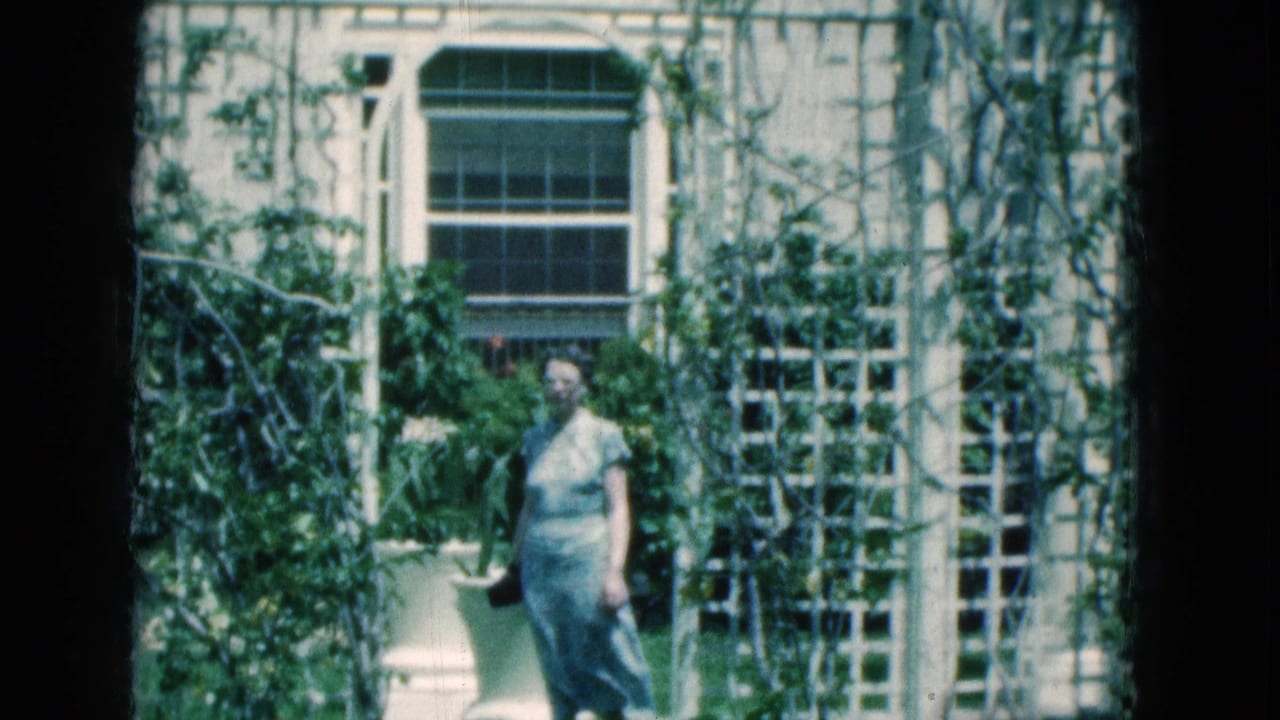 In 1975 Florida home-movie footage, a senior woman carries a dark purse while walking past white trellises and large planters outside a vintage building.