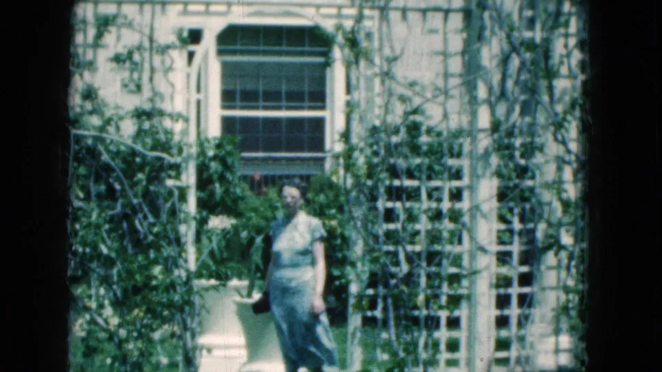 In 1975 Florida home-movie footage, a senior woman carries a dark purse while walking past white trellises and large planters outside a vintage building.