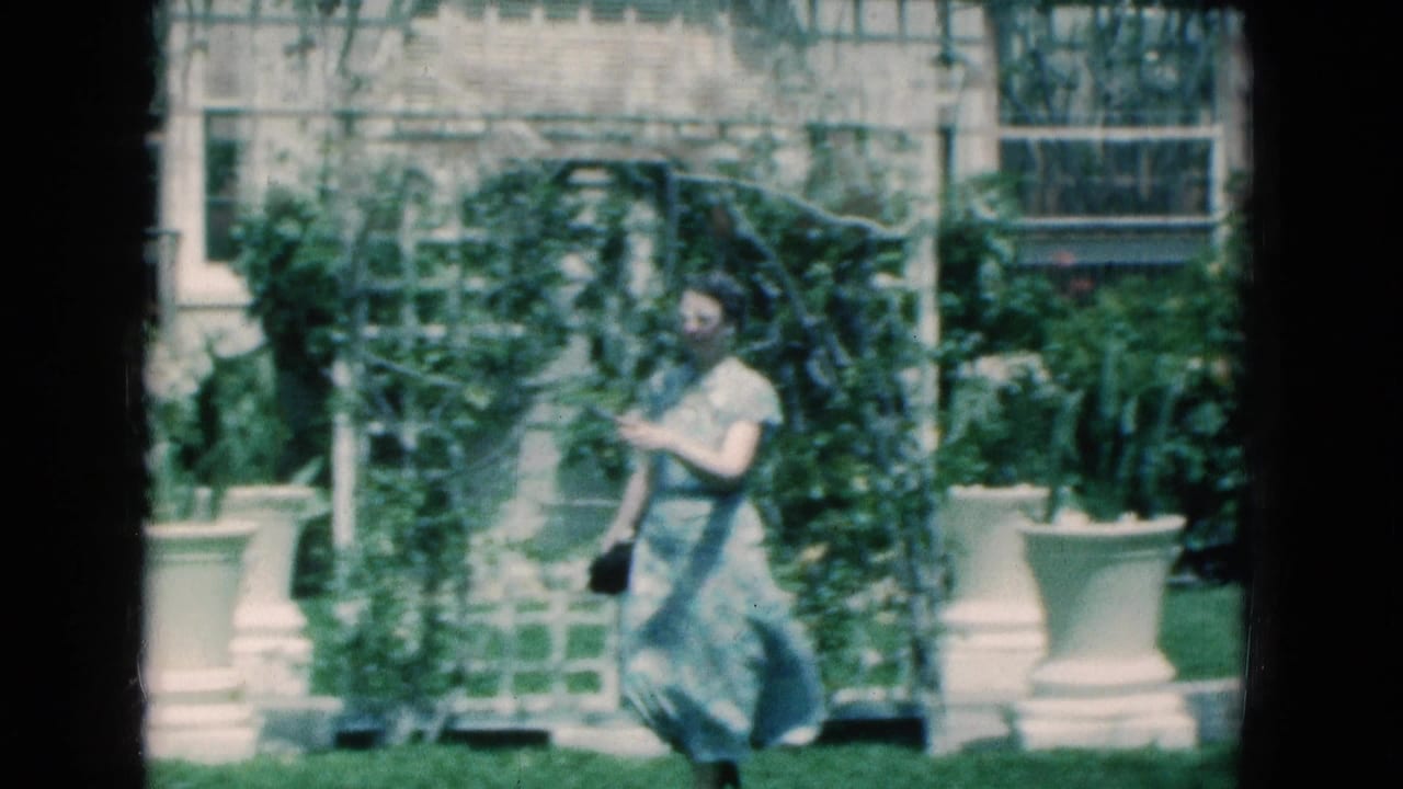Senior woman walks across grass near a trellis, planters, and a light-colored building.