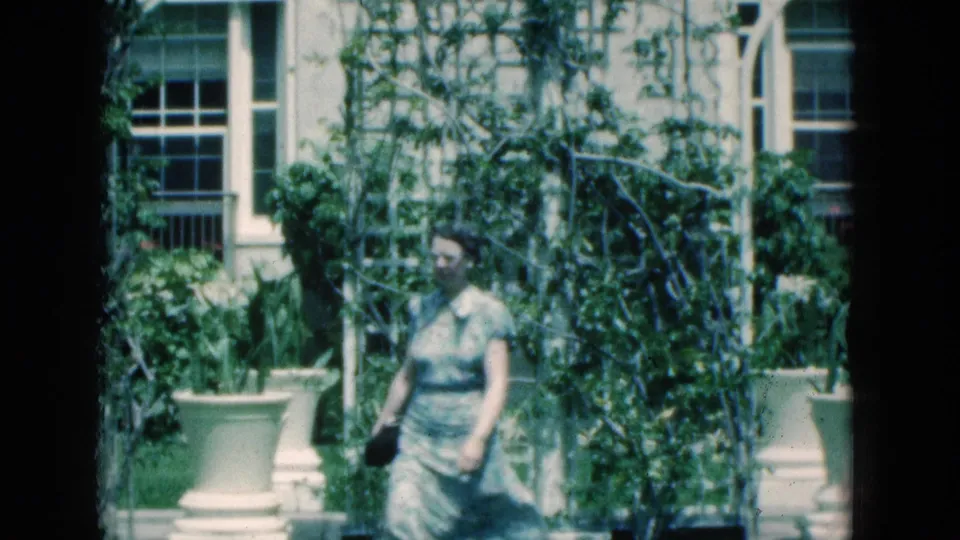 The woman appears closer in frame, walking with a small dark purse in a landscaped 1975 Florida exterior with climbing vines and building windows behind her.