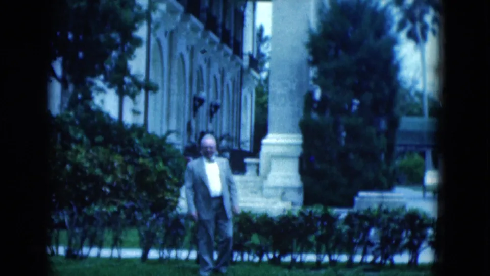 A senior man in a suit walks along a landscaped exterior in Florida, with hedges, trees, columns, and an older building facade visible behind him.
