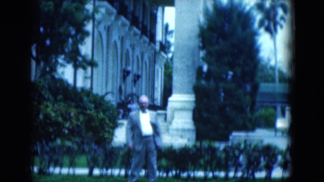 Man in a light suit stands on a lawn near hedges and a columned building.